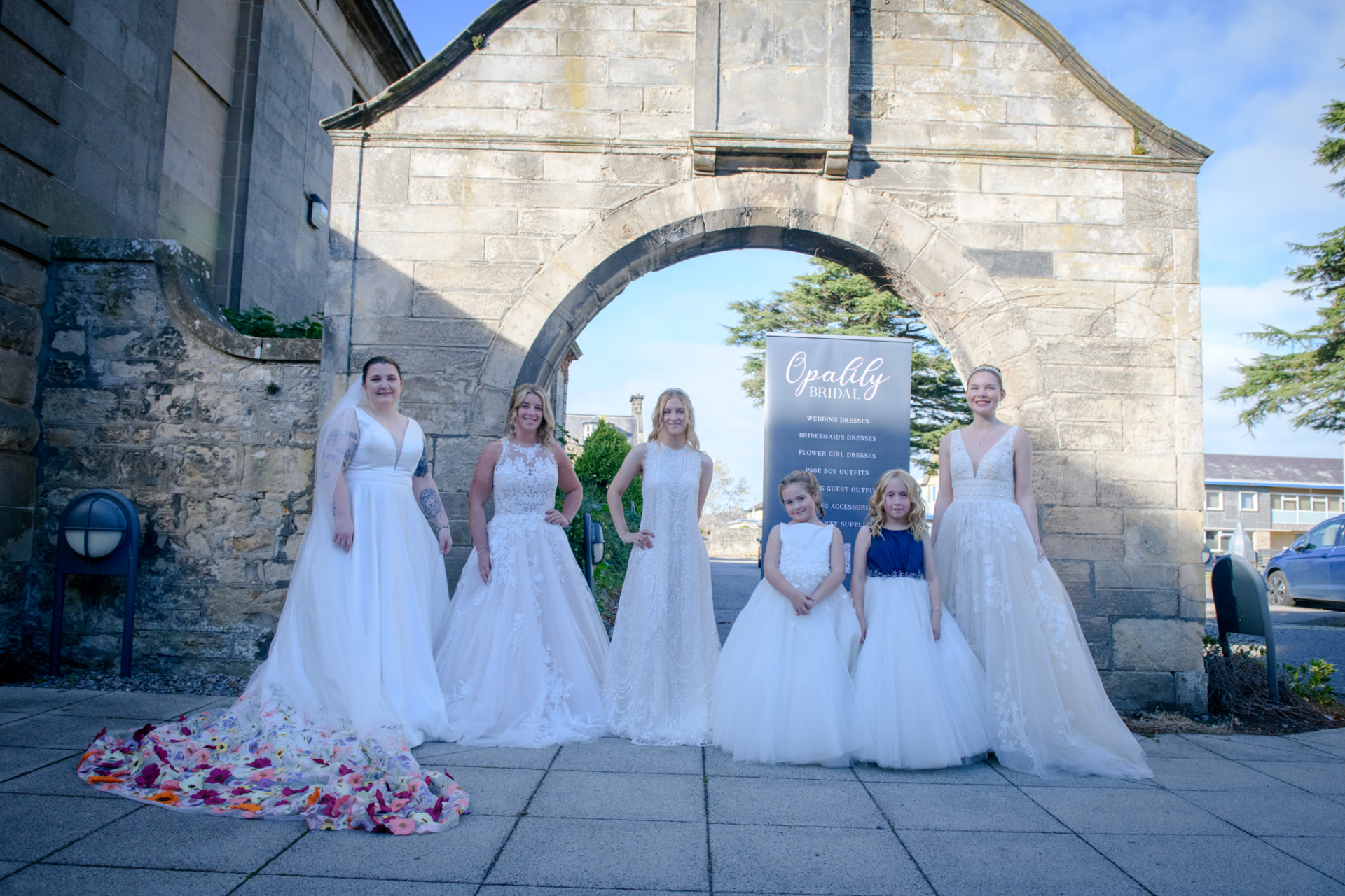 Brides and flower girls in gowns beneath the stone arch at Moray College UHI, Elgin
