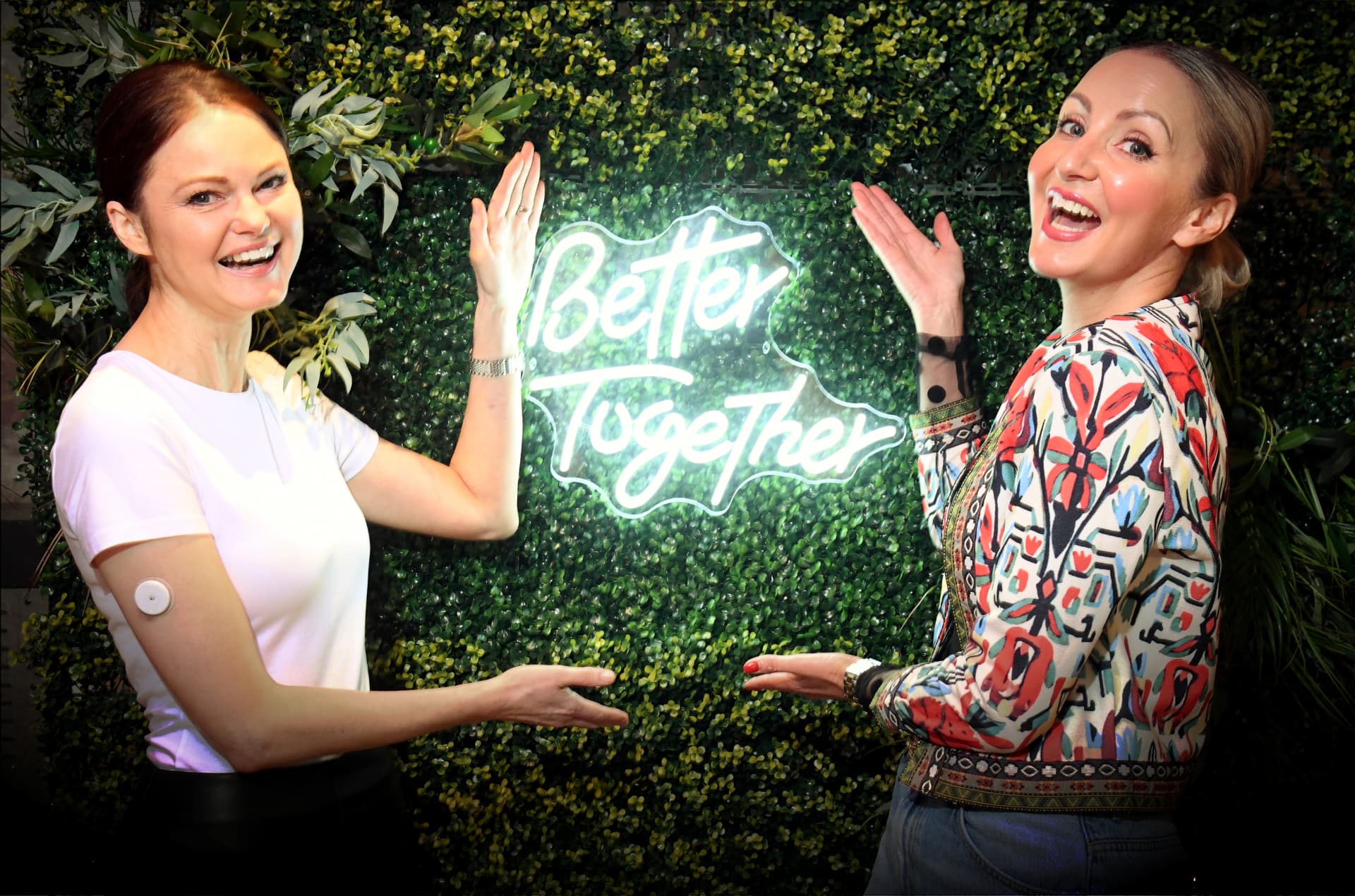 Two women beside a 'Better Together' neon sign on a foliage wall