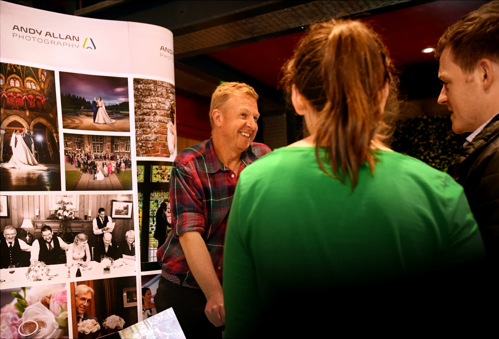 A wedding photographer chatting with a couple beside his display banner