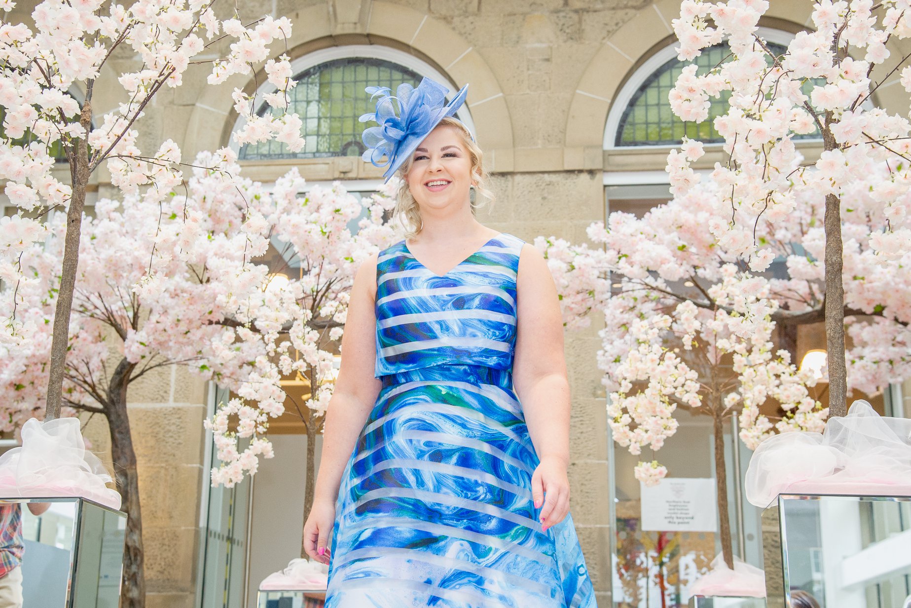 Model in a blue printed gown and fascinator among blossom trees at the Moray Wedding Show